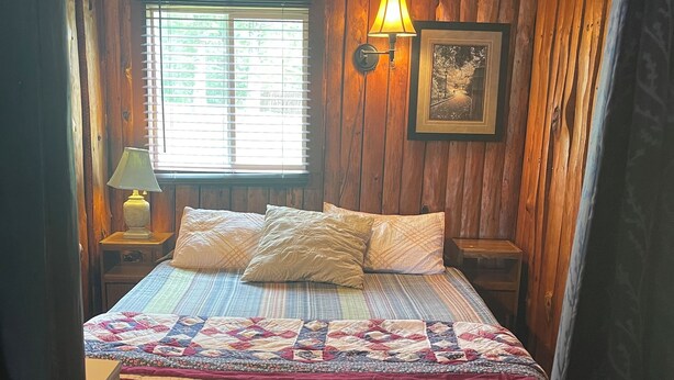 A wooden cabin bedroom with a bed, a nightstand, a lamp, a window, and a wall-mounted picture. - Cozy cabin on Island Lake (Winter)