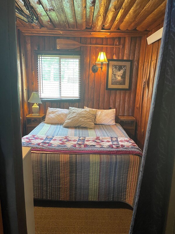 A wooden cabin bedroom with a bed, a nightstand, a lamp, a window, and a wall-mounted picture.