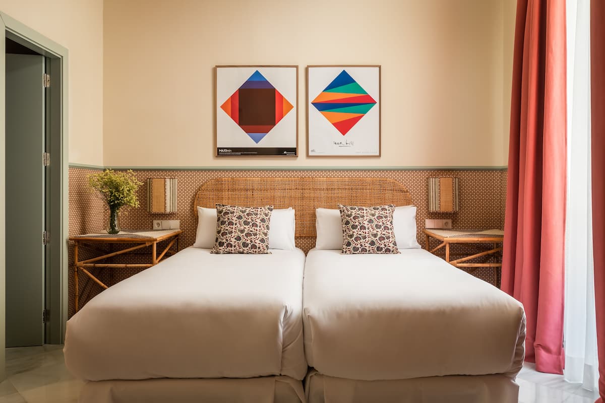 Habitación de hotel con dos camas, un cabecero de madera, una mesita con un florero y dos cuadros en la pared.