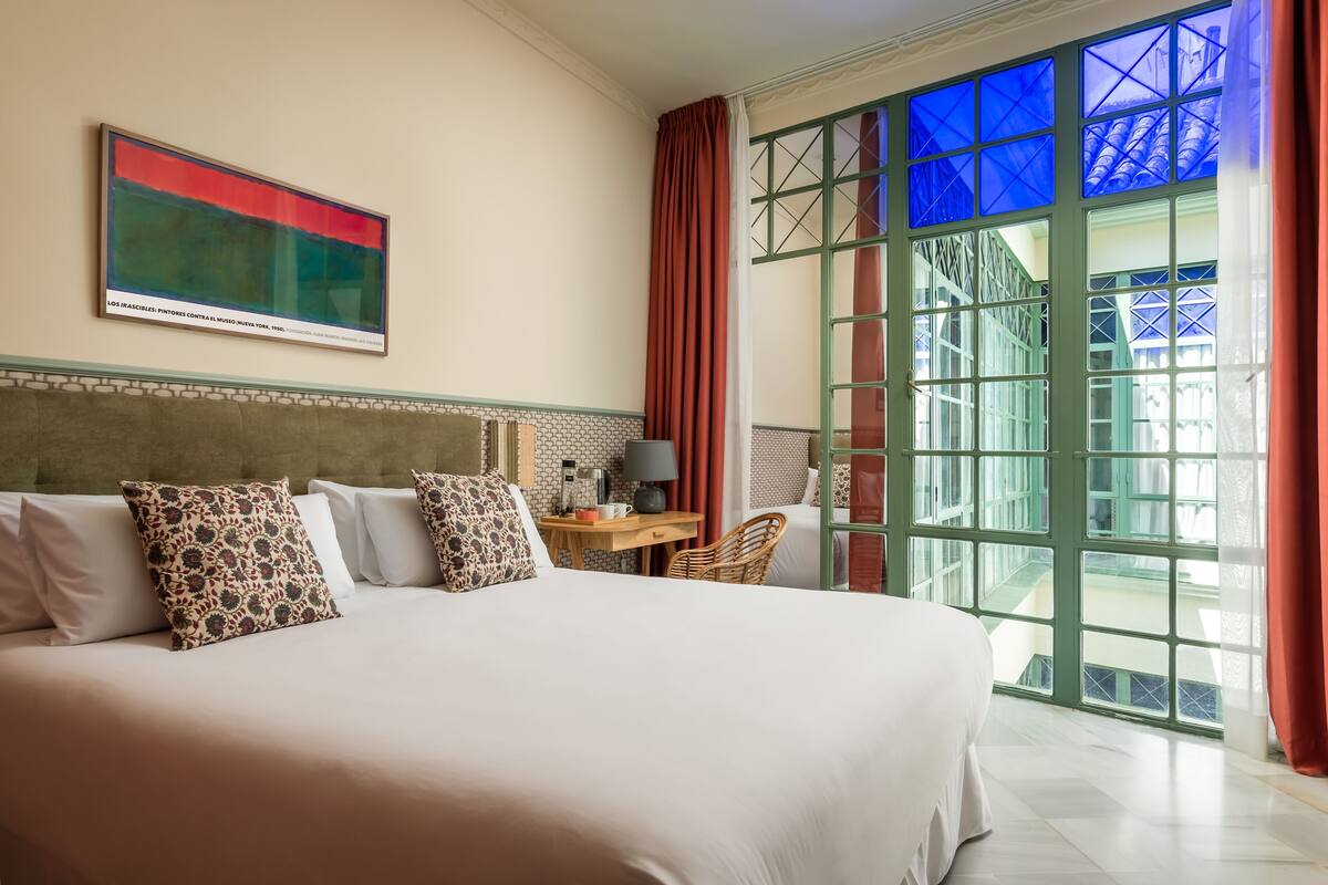Una habitación de hotel con una cama grande, un escritorio con lámpara y vistas a través de una ventana con marco verde.