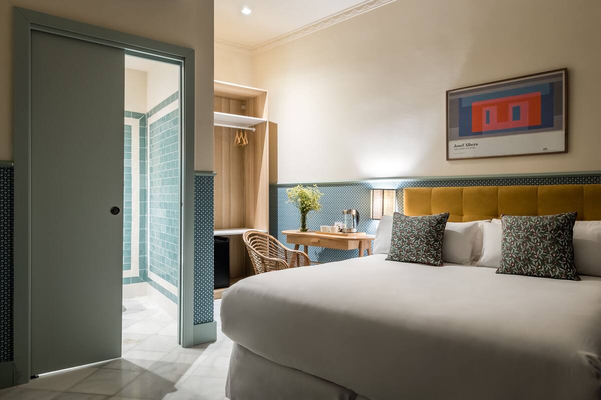 Habitación de hotel con cama, escritorio y silla. Se observa una obra de arte enmarcada en la pared y una puerta que da a otra habitación.