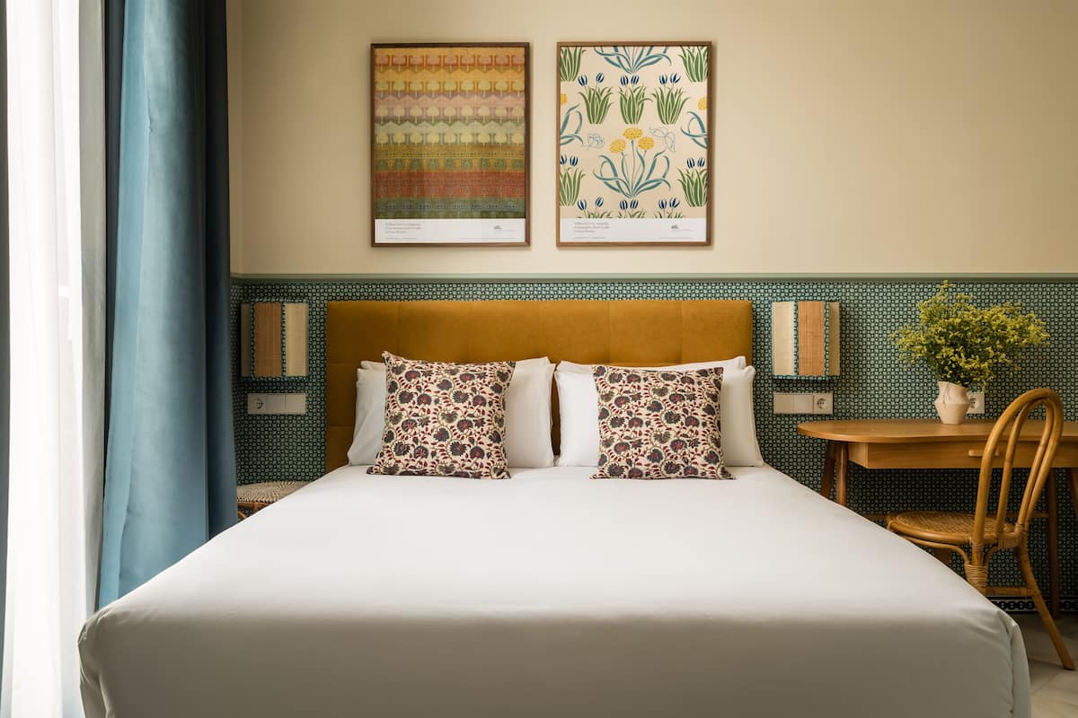Una cama bien hecha con almohadillas estampadas, un cabecero de madera y una mesita con una silla.