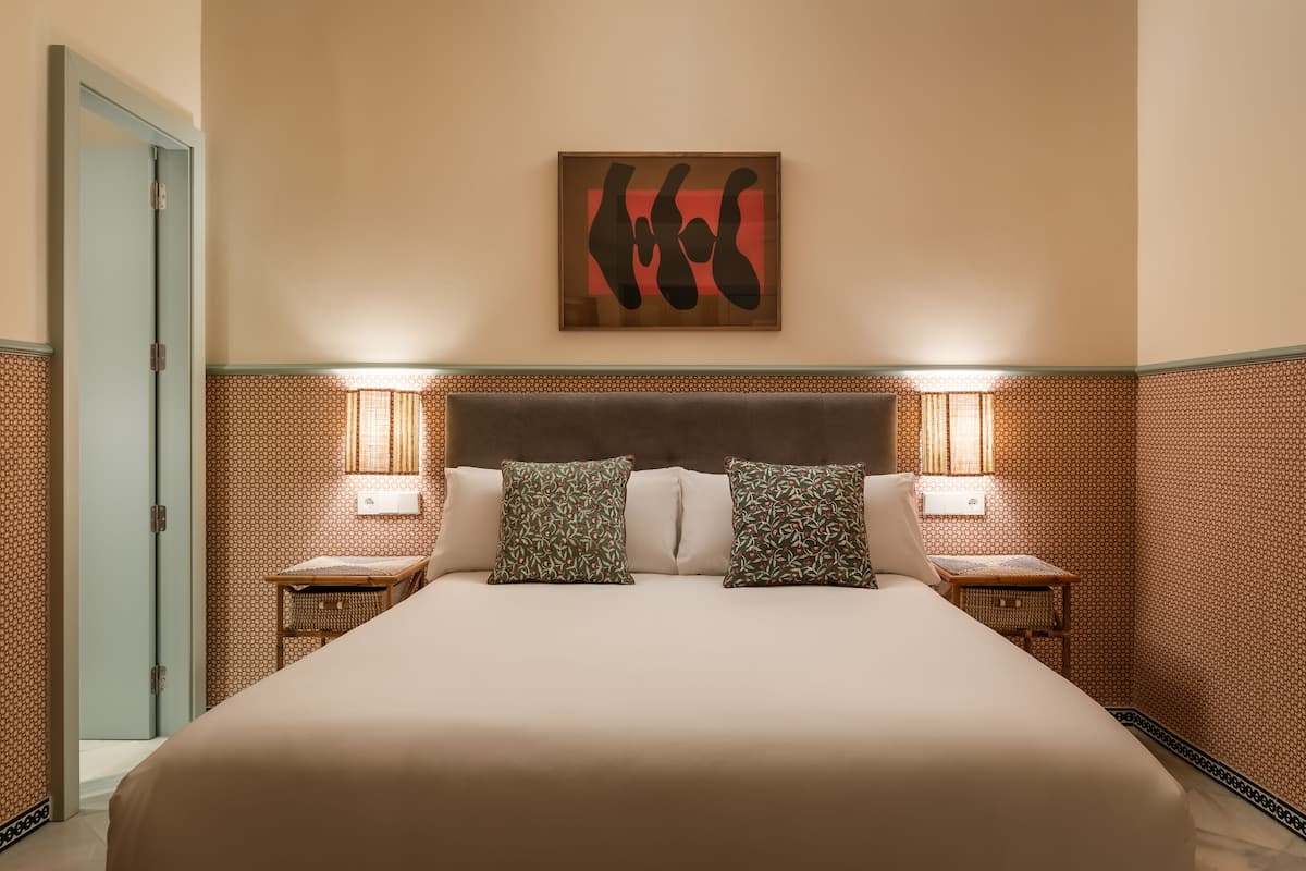 Una habitación de hotel con una cama grande, dos mesitas de noche con lámparas, una obra de arte colgada en la pared y papel tapiz estampado.