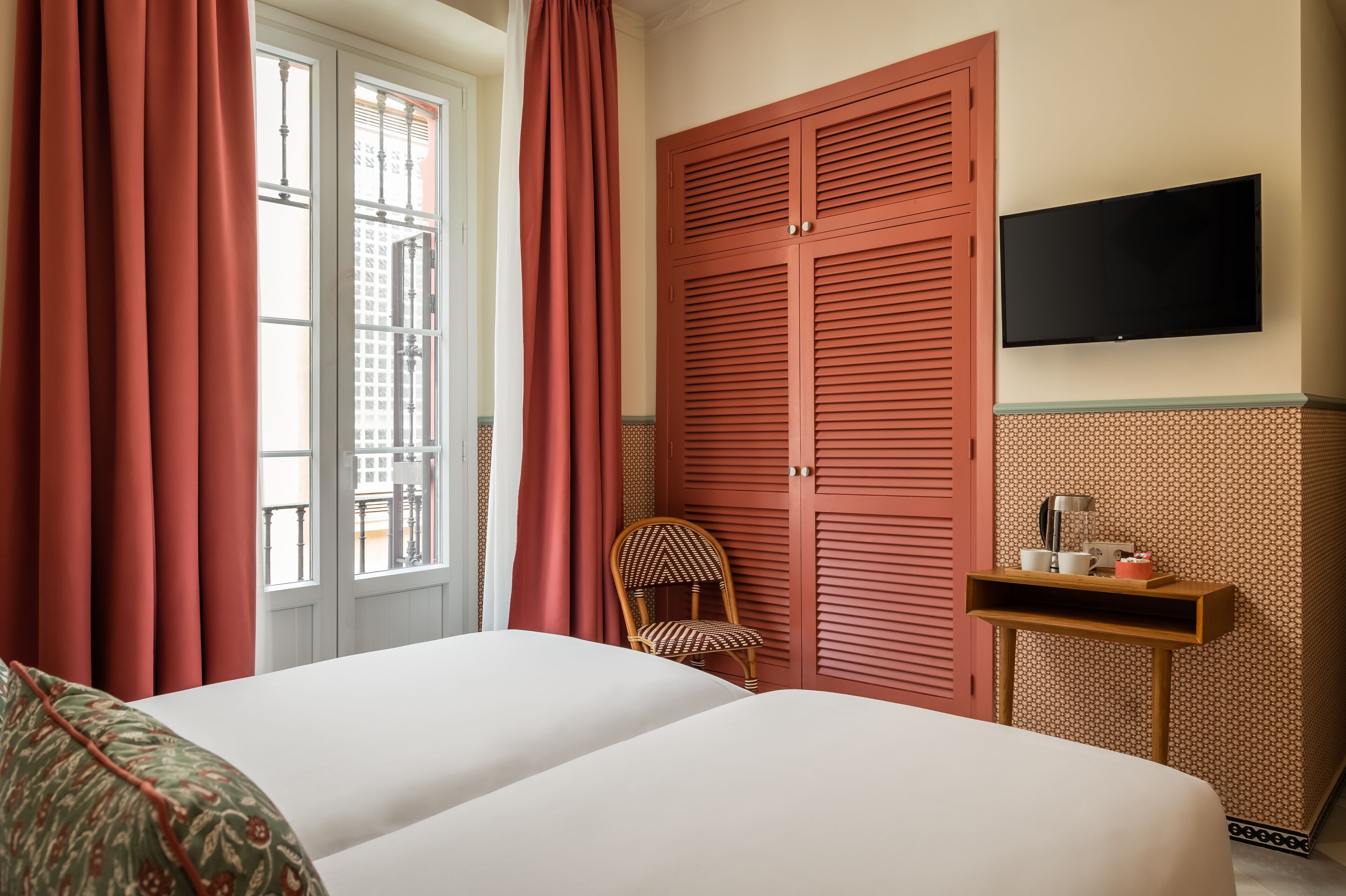 A hotel room with a bed, a chair, a TV, and a small table with a lamp and a cup.