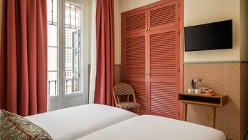 A hotel room with a bed, a chair, a TV, and a small table with a lamp and a cup.