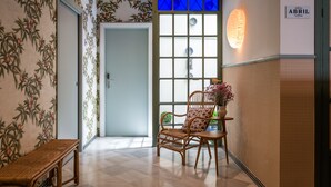 Une pièce dotée d’une grande fenêtre, d’un fauteuil en osier, d’une petite table sur laquelle est posé un vase, et d’un mur orné d’un motif floral.