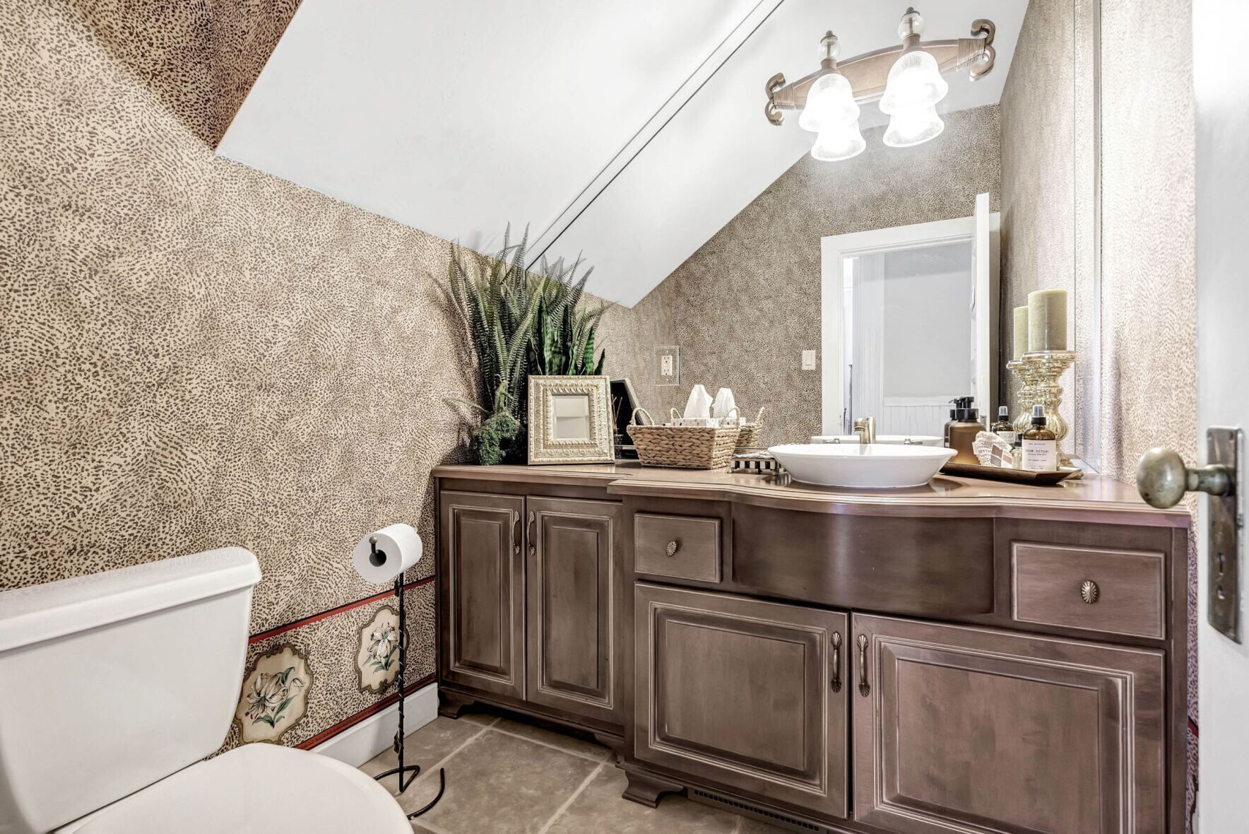 Une salle de bain avec un grand miroir, un lavabo et un meuble. On peut voir une plante sur le comptoir et des toilettes dans un coin.