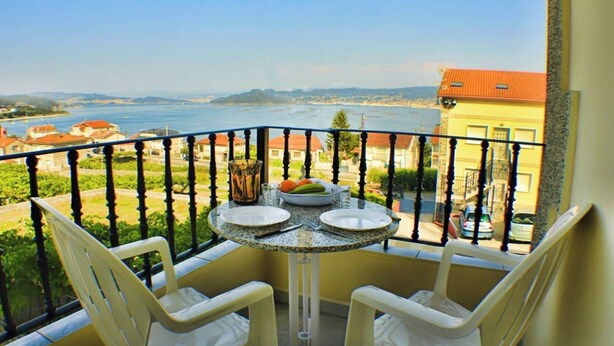 A balcony with a table set for two, overlooking a coastal view with buildings and a body of water. - Apartment - Standard Floor - Without Terrace (Raxó)