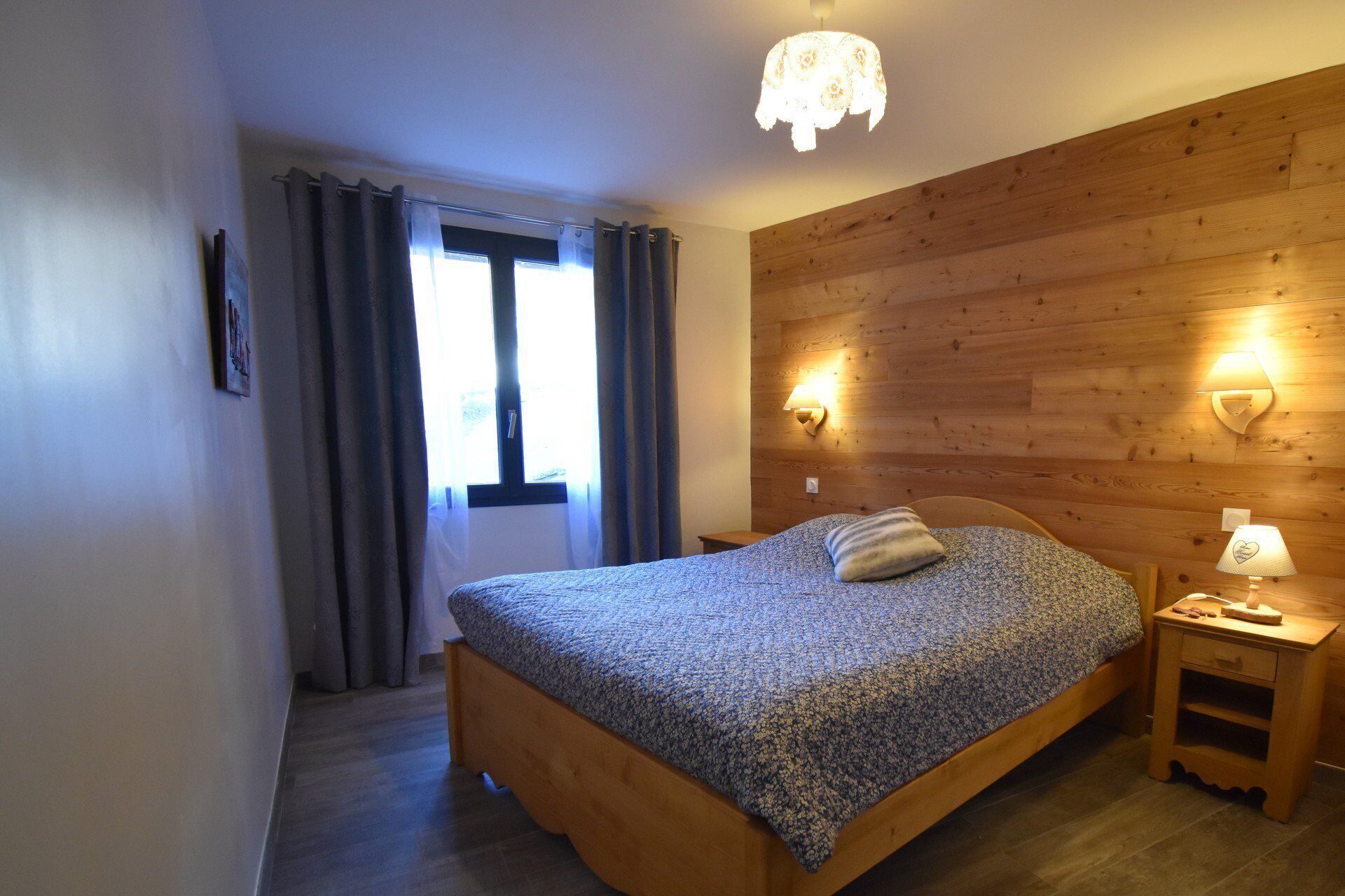 A bedroom with a wooden bed, a nightstand with a lamp, a window with curtains, and a wooden wall panel.
