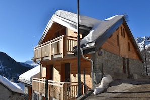 A wooden chalet with a balcony, stone walls, and a snow-covered roof.