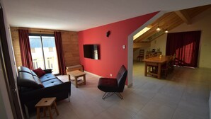 A living room with a red wall, a flat-screen TV, a sofa, a coffee table, and a dining area with a table and chairs.