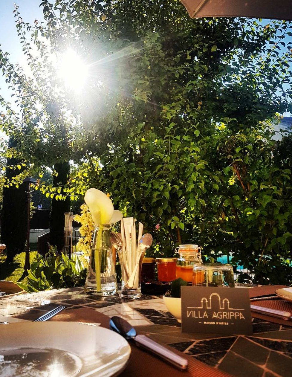 Un espace de restauration en plein air avec une table dressée pour deux personnes, une enseigne indiquant « Villa Agrippa », et un décor de verdure luxuriante.