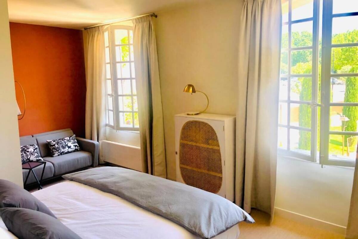 Un dormitorio con cama, sofá, radiador y ventanas amplias con cortinas.