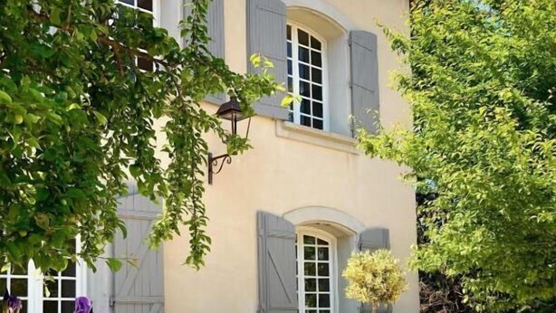 A two-story building with a light-colored facade, grey shutters, and a tiled roof. There are steps leading up to the entrance, and purple flowers in pots are visible in the foreground. - Villa Agrippa (Orange)