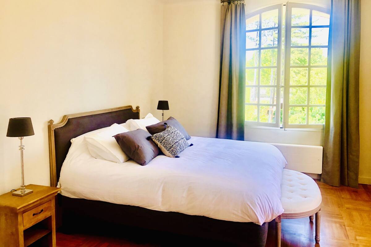 Una cama bien hecha con sábanas blancas y varias almohadas, una lámpara de noche y una ventana con cortinas.