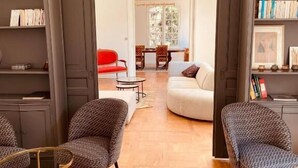 A modern living room with a wooden parquet floor, a white sofa, two patterned armchairs, a red chair, and a dining area in the background. - Villa Agrippa (Orange)