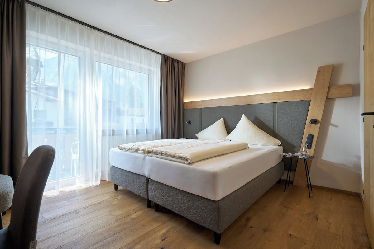 A modern hotel room with a large bed, a wooden headboard, a small table with a lamp, and a view of the outdoors through sheer curtains.