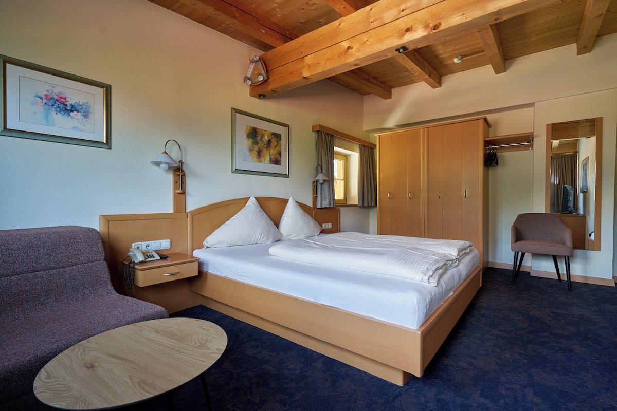 A hotel room with a wooden bed, a sofa, a small table, a wardrobe, and two framed artworks on the wall.