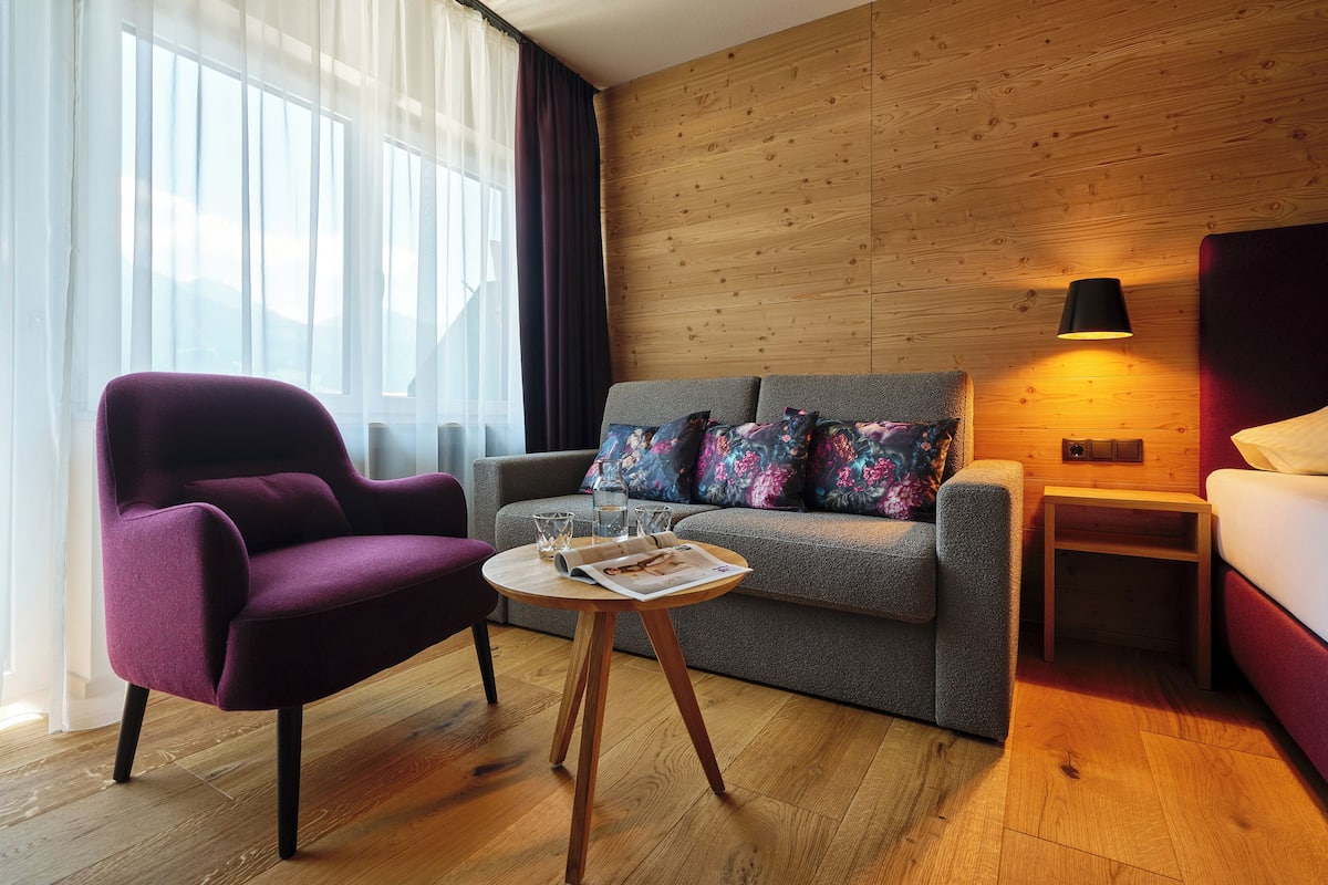 A modern hotel room with a wooden wall, a grey sofa with colorful pillows, a purple armchair, a small wooden table with a magazine, and a bedside lamp.