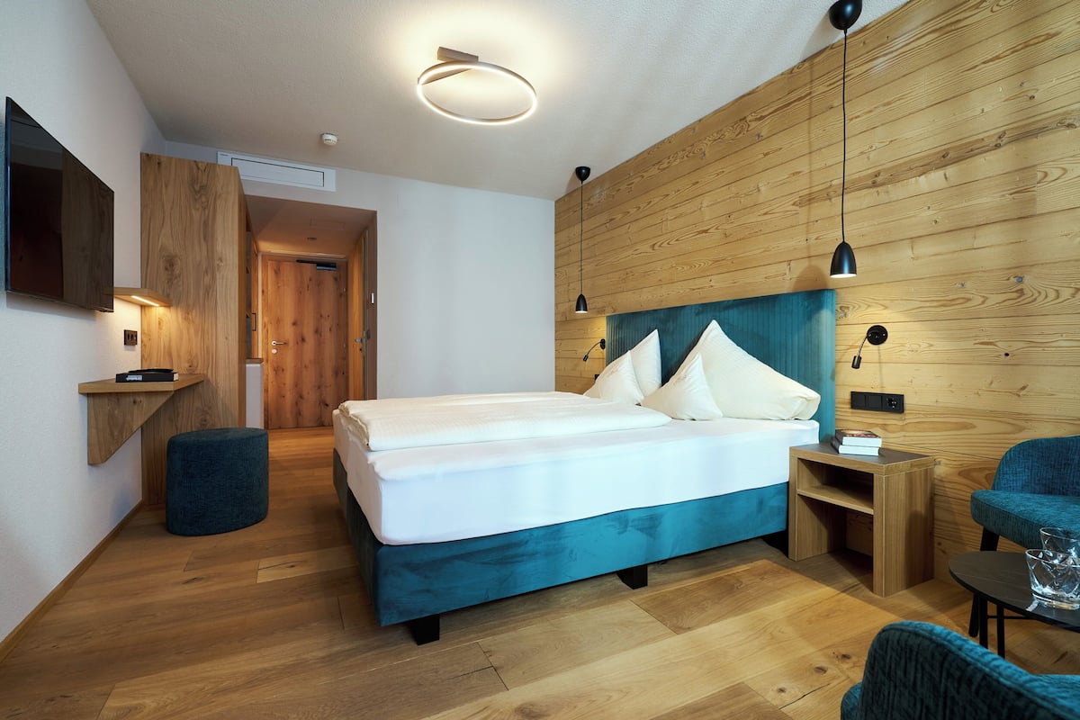 A modern bedroom with a wooden headboard, a bed with white linens, a small bedside table, a chair, and a wooden floor.