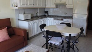 Una cocina compacta con armarios blancos, microondas, cafetera y refrigerador. Hay una mesa de comedor con sillas y un sofá en la zona de estar.