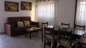 Una sala de estar con un sofá marrón, una mesa de centro y una mesa de comedor con sillas. Hay dos cuadros en la pared y una ventana con cortinas.