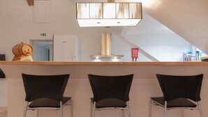 Een bar met drie zwarte stoelen, een houten toog, een moderne lamp en een witte koelkast op de achtergrond.