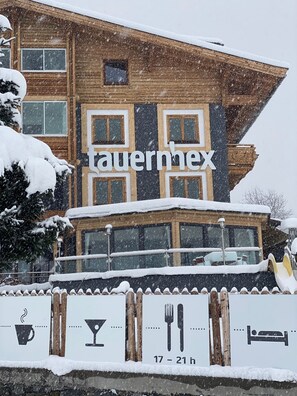 A wooden chalet with a balcony, snow-covered roof, and a sign indicating a 17-21 hour stay. - Hotel Restaurant tauernhex (Piesendorf)