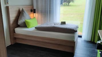 A small, modern room with a bed, a bedside table, a window with curtains, and a view of the outdoors.