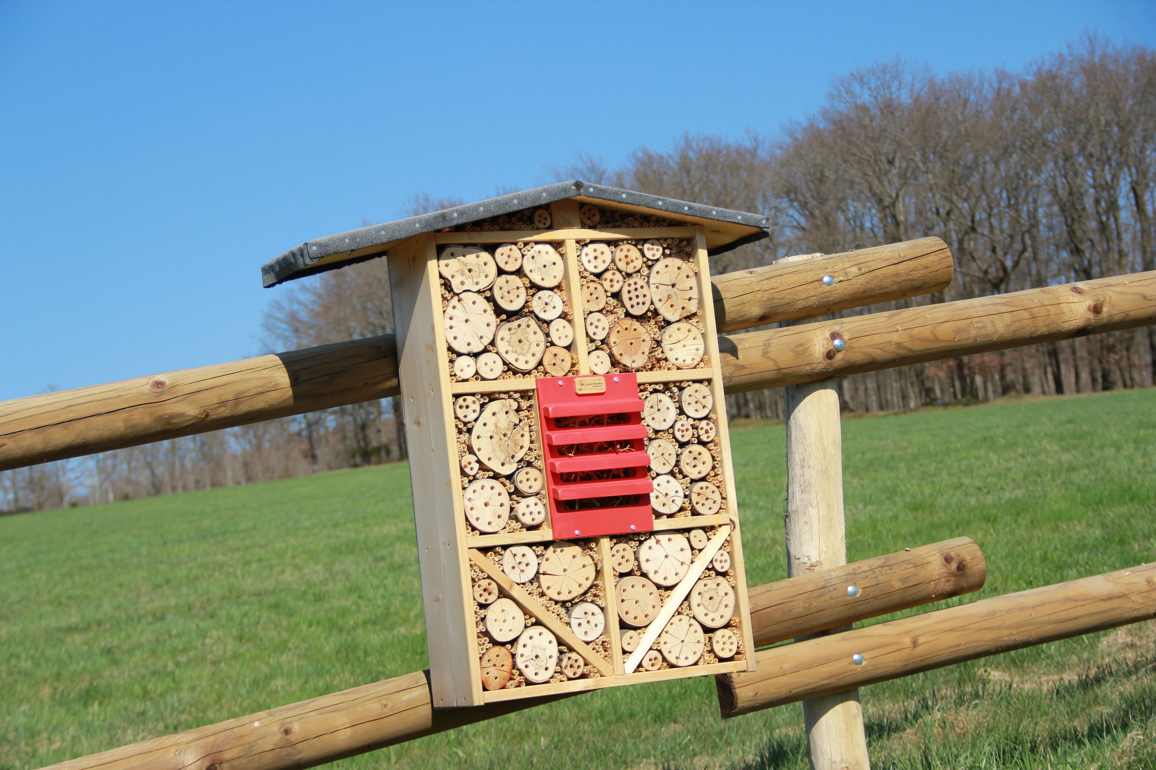 Una estructura de colmena de madera con un panel de ventilación rojo, unida a una valla de madera en un campo cubierto de hierba.