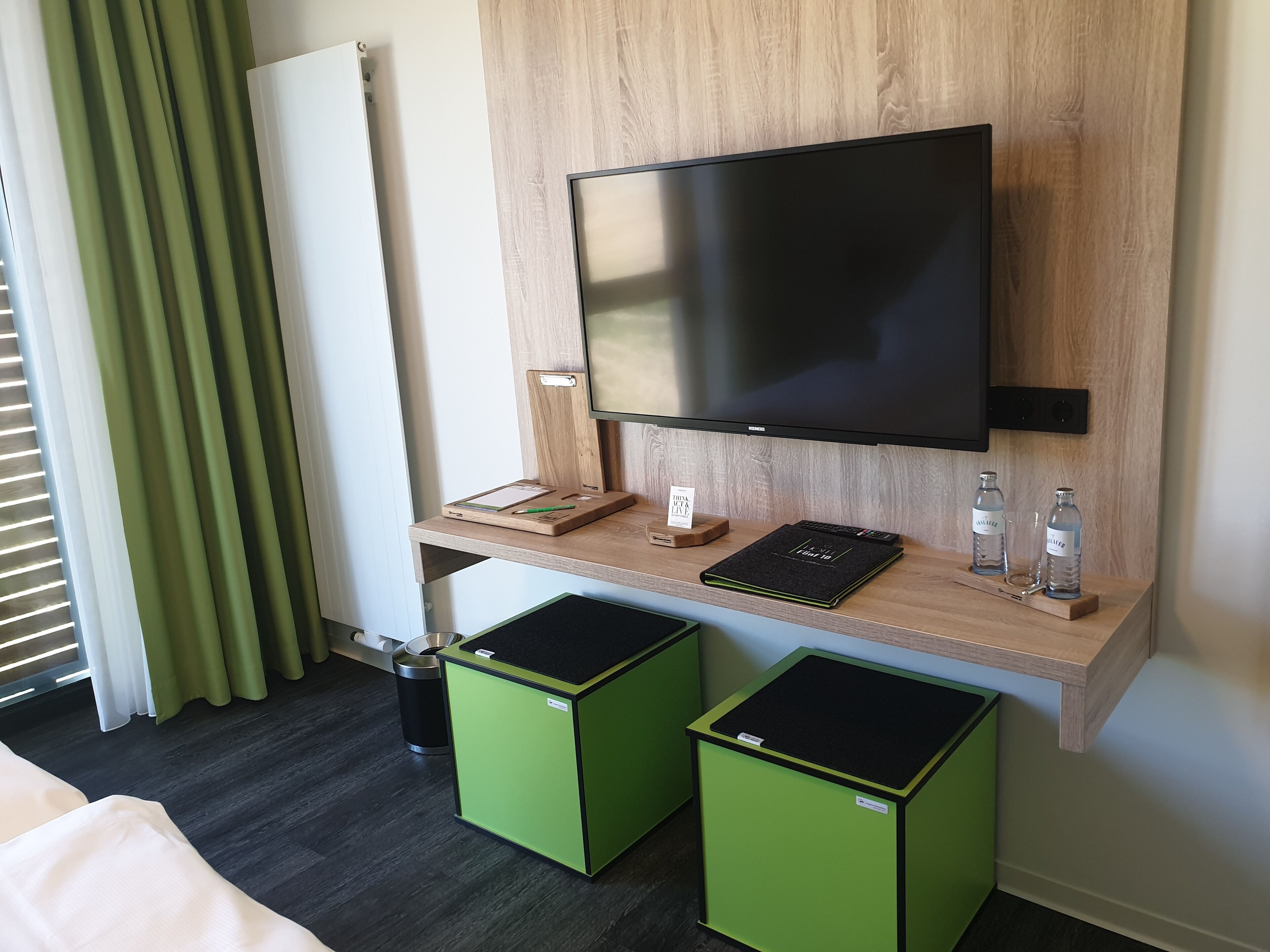 A hotel room with a wooden desk, a flat-screen TV, a small trash bin, and two green waste bins.