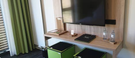 Habitación de hotel con un escritorio de madera, un televisor de pantalla plana, un bote de basura pequeño y dos cubos verdes para residuos.