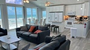 Una sala de estar moderna con un amplio ventanal con vista al océano, una cocina con gabinetes blancos y un comedor con taburetes de bar.