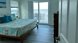 Un dormitorio con cama, ventilador de techo, vista al océano a través de una puerta corrediza de vidrio y un aparador de madera.