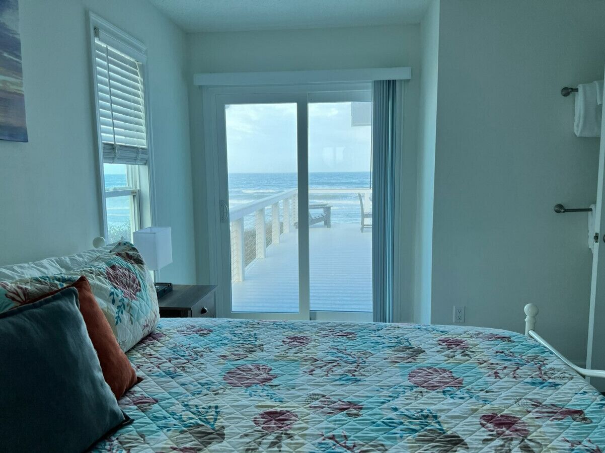 Een slaapkamer met een bed, een nachtkastje, een raam met zonwering en uitzicht op de oceaan door een schuifdeur.