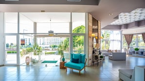 A spacious living area with a teal sofa, a coffee table, and a dining area with a chandelier. Large windows provide a view of the outdoors. - Happy Days Hotel (Rhodes)