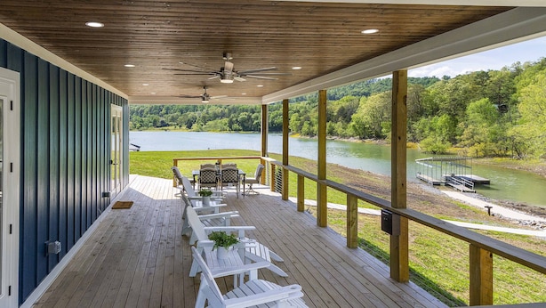 Eine überdachte Terrasse mit weißen Adirondack-Stühlen und einem Tisch, mit Blick auf einen See mit einer Anlegestelle und einem Boot.