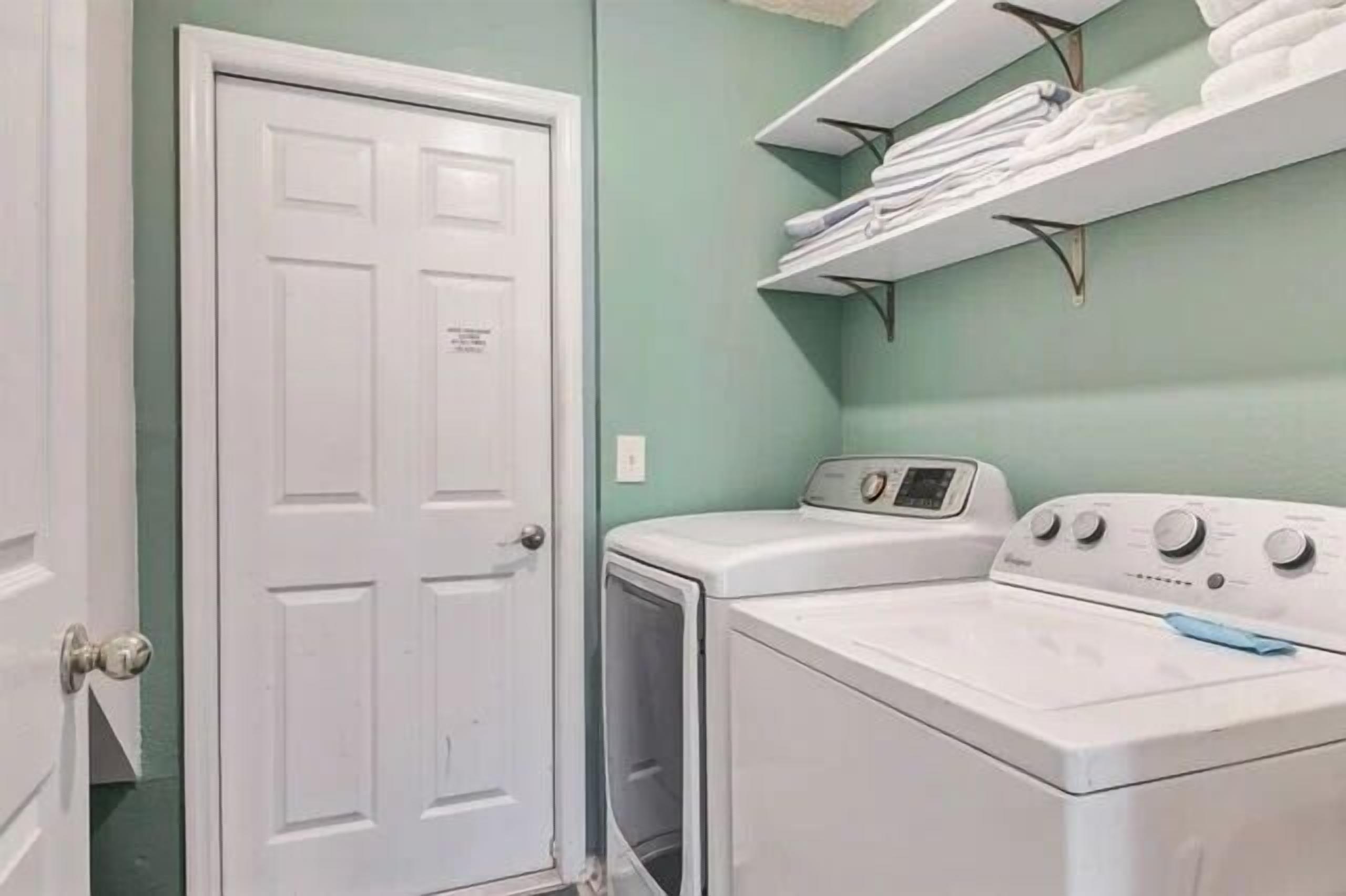 Une buanderie avec une porte blanche, un lave-linge et un sèche-linge. Des étagères au-dessus du sèche-linge servent à ranger les serviettes pliées.