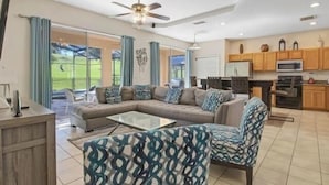A spacious living room with a large sectional sofa, a coffee table, and patterned armchairs. A ceiling fan and recessed lighting are visible.