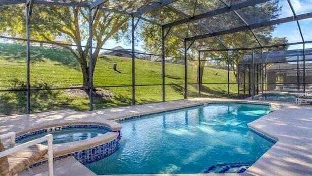 A pool with a hot tub, surrounded by a glass enclosure, with a view of a grassy area and trees.