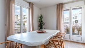 A dining area with a large table, four wooden chairs, a potted plant, and large windows with curtains. - LOFUS bio-suites (Athens)