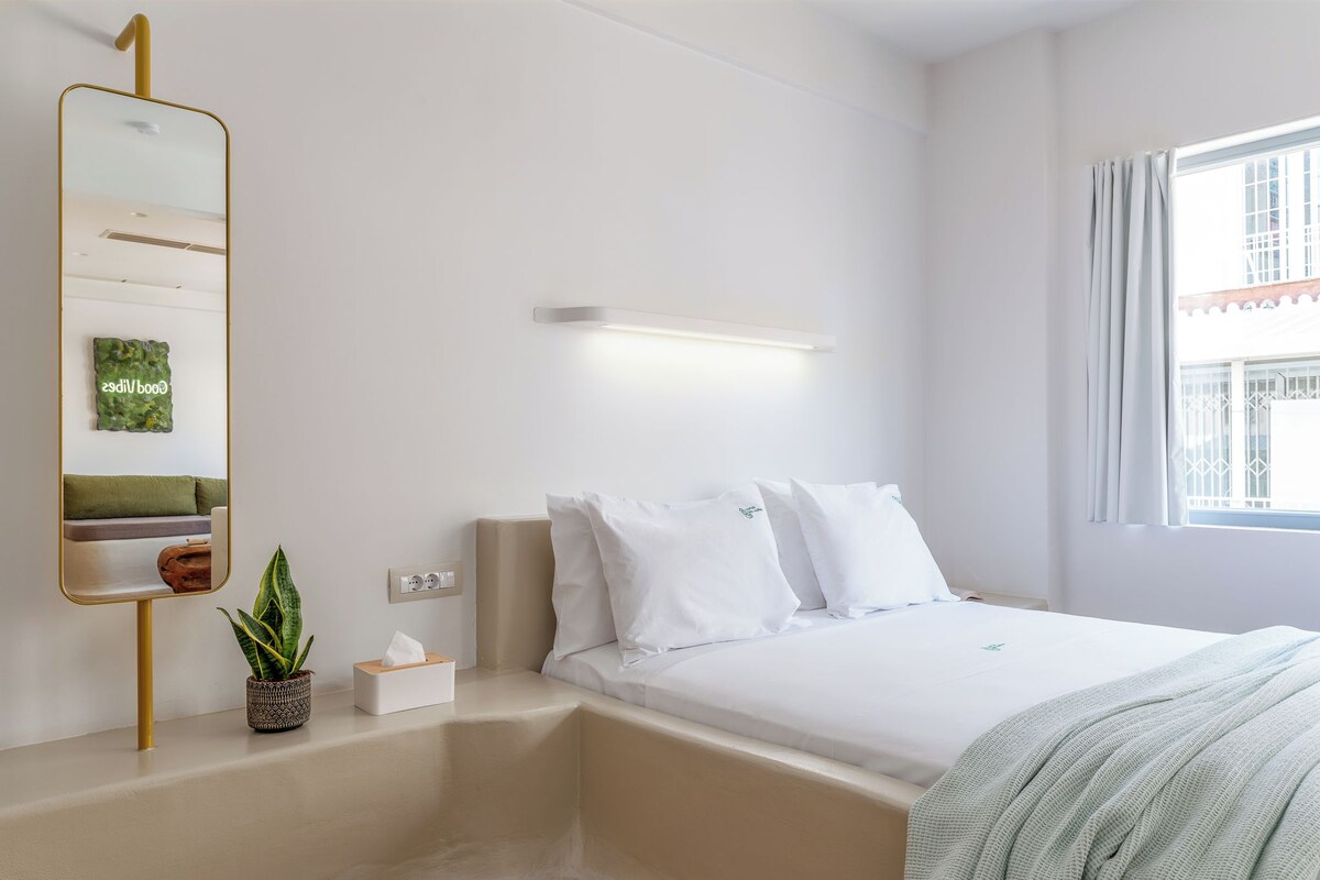 A hotel room with a bed, a bedside table with a tissue box, a potted plant, a mirror, and a window with curtains.