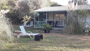 A small, single-story building with a porch, a covered seating area, and a lawn with a lounge chair and a speaker.