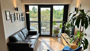 Una sala de estar moderna con un sofá de cuero negro, un sillón azul y una mesa de centro de madera. Se observan varias plantas en macetas y cuadros enmarcados en las paredes.