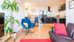 Una sala de estar moderna con un sofá de cuero negro, un sillón azul y una alfombra roja. Una planta grande está en la esquina, y un televisor de pantalla plana sobre un pedestal de madera.