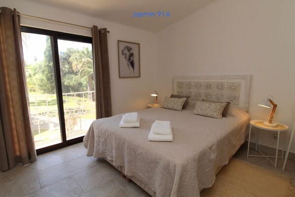 A bedroom with a large bed, two bedside tables, a view of greenery through a sliding door, and a framed artwork on the wall.