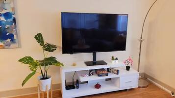 A modern living room with a flat-screen TV on a white entertainment unit, a potted plant, and a small table with decorative items.