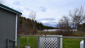 A deck with a hanging planter, a white lattice fence, and a view of a grassy area with trees and a body of water in the background. - Quiet and Cozy Seaside Cottage with Amazing Sunsets (Steuben)