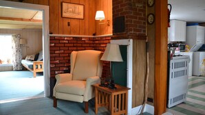 A cozy room with a brick fireplace, a comfortable armchair, a small table with a lamp, and a view into another room with a bed and curtains. - Quiet and Cozy Seaside Cottage with Amazing Sunsets (Steuben)