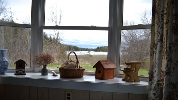 A window with a view of a lake and trees, a wooden shelf with a basket, a birdhouse, and a vase. - Quiet and Cozy Seaside Cottage with Amazing Sunsets (Steuben)